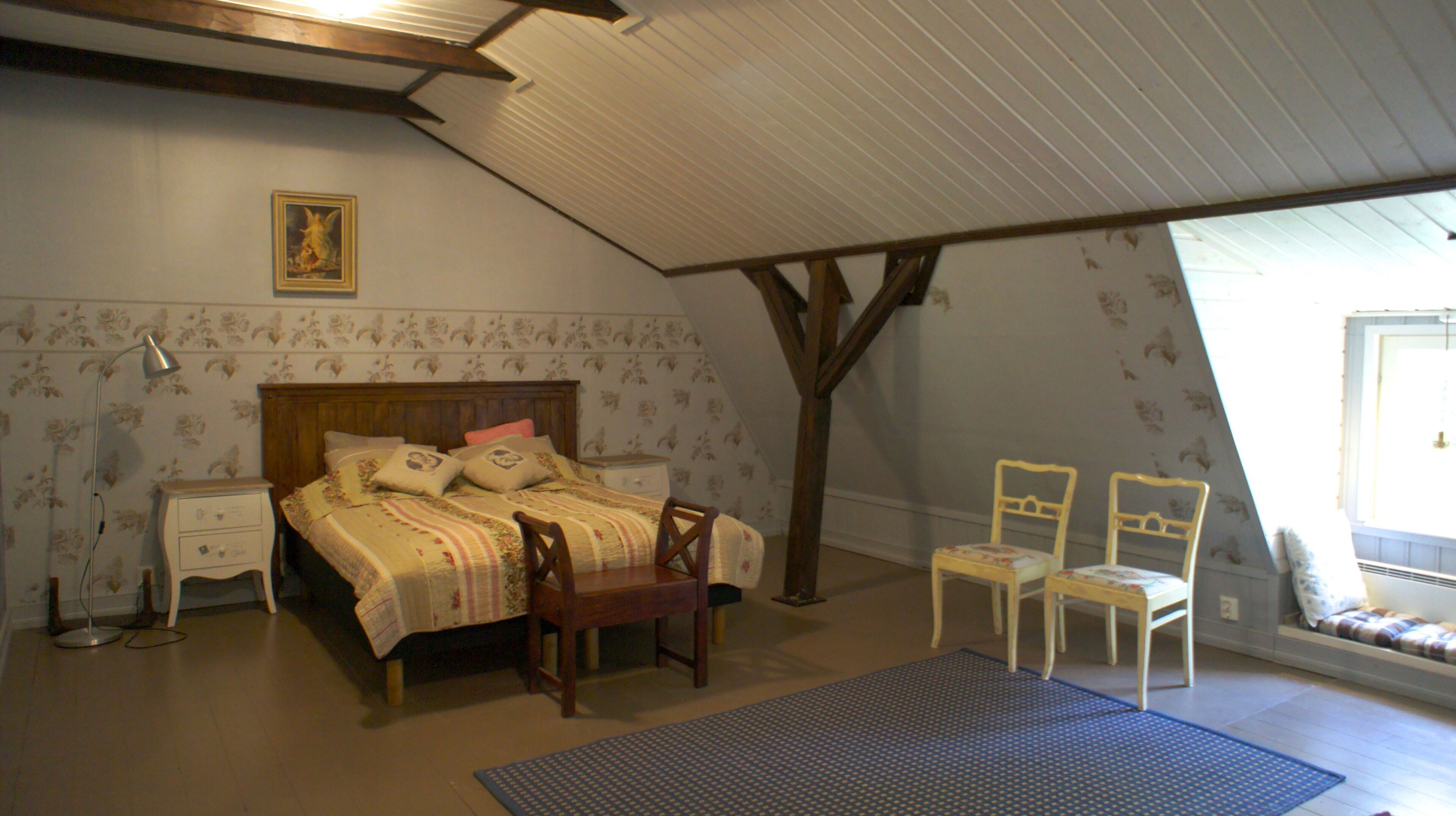 Een slaapkamer met een schuine kap, een bed met bloemenbeddengoed, twee stoelen, een nachtkastje en een lamp.