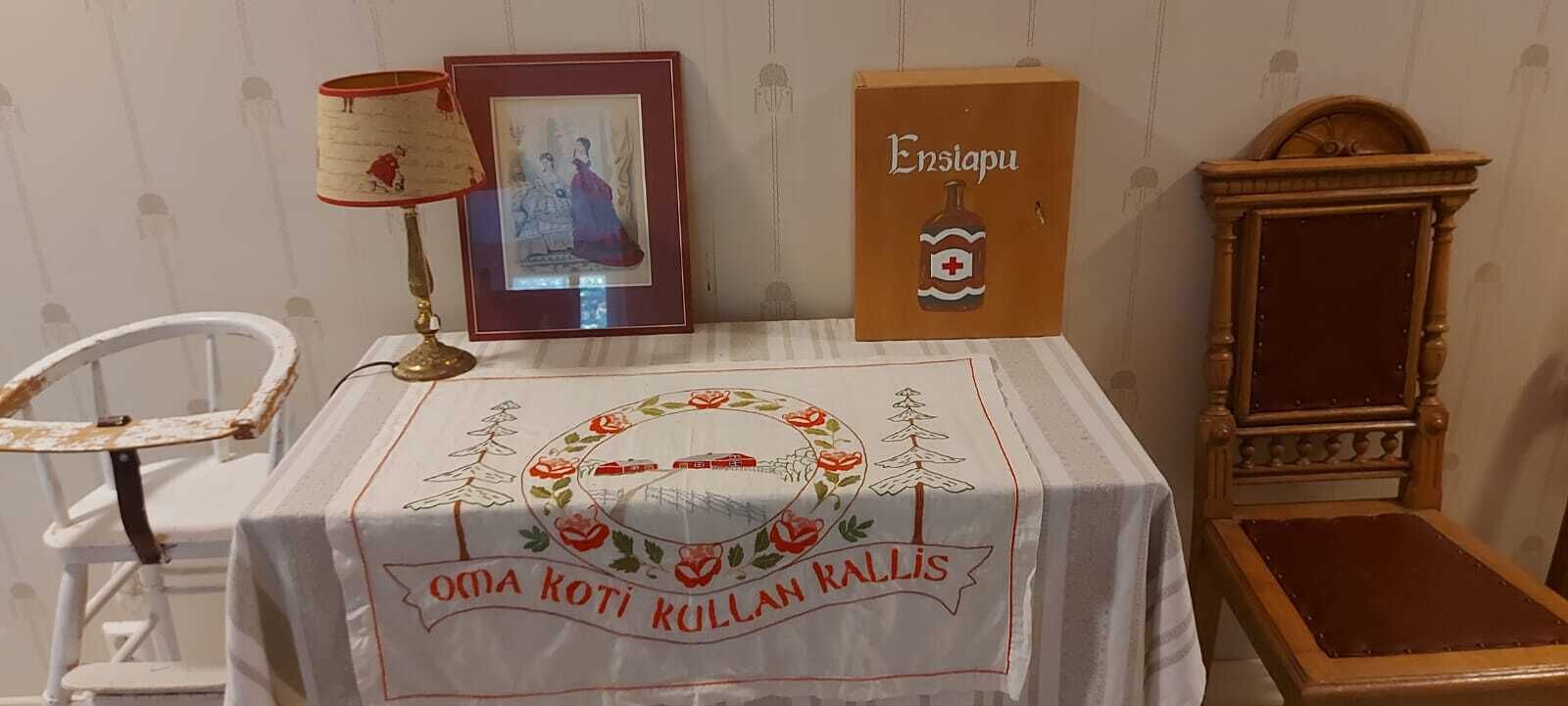 Una habitación con una mesa cubierta por un mantel con diseño floral y texto, un cuadro enmarcado, una lámpara, un mueble de madera y una silla.