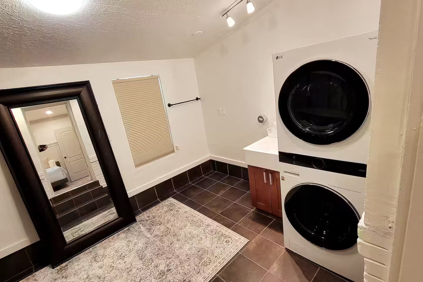A laundry room with two washing machines, a mirror, a window with blinds, and a patterned rug.
