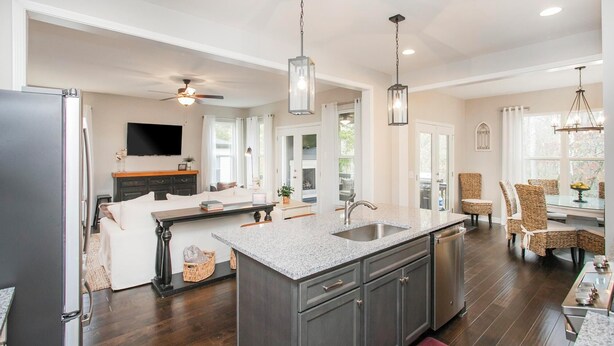 A modern kitchen with dark cabinetry, a granite countertop, and a central island. There is a dining area with wicker chairs and a chandelier.