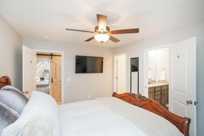 Une chambre à coucher avec un lit, un ventilateur de plafond, une télévision et une salle de bain visible à travers une porte ouverte.