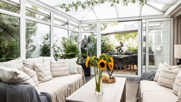 A conservatory with a sofa, a coffee table with a vase of sunflowers, and a view of the garden outside.