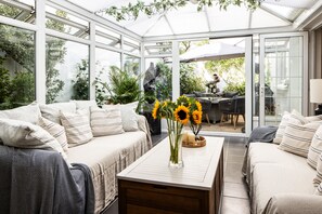 A conservatory with a sofa, a coffee table with a vase of sunflowers, and a view of the garden outside.
