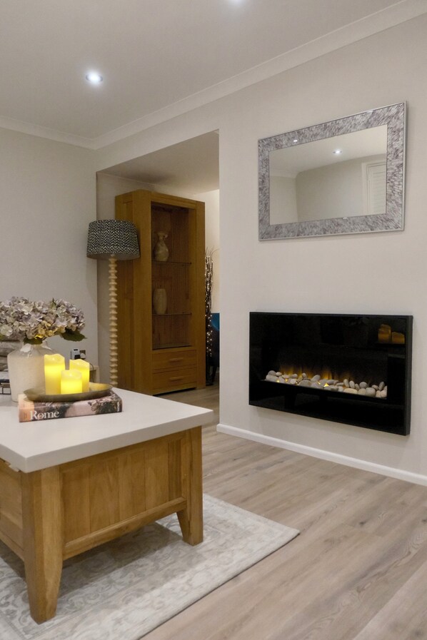 A modern living room with a white table, candles, a book, a fireplace, and a mirror on the wall.