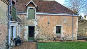 A rustic building with a brick exterior, a green door, and a small window. There is a grassy area in front and a bench to the left.
