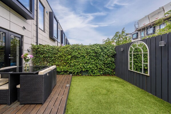 A modern outdoor patio with wicker furniture and a small table, a neatly trimmed lawn, and a black fence with a window.