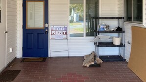Un porche delantero con una puerta azul, un cartel de 'For Rent', una ventana y un estante con objetos.