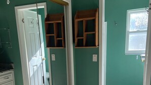 Una habitación con paredes verde azuladas, suelo de madera y una puerta blanca. Se observan dos estantes de madera montados en la pared, y un armario blanco a la izquierda.