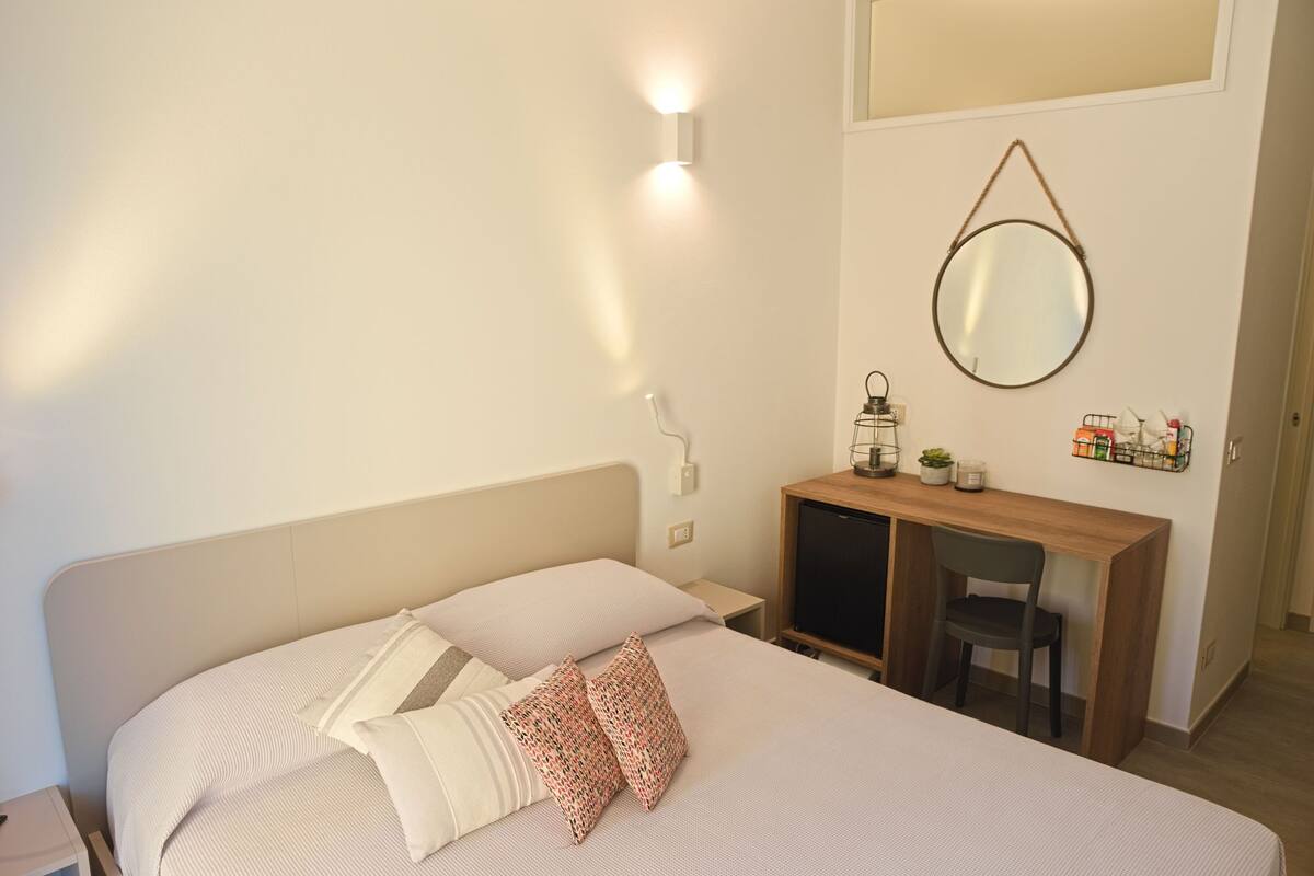 Ein ordentlich eingerichtetes Schlafzimmer mit Bett, Nachttisch, Schreibtisch, Stuhl und einem Spiegel an der Wand.