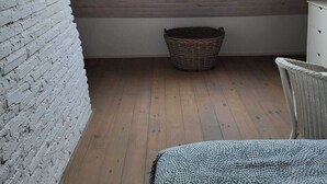 Una stanza con soffitto spiovente, una finestra con tende, un letto, un cesto di vimini e un muro in mattoni bianchi.