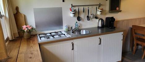 A compact kitchen with a gas stove, a sink, and a coffee maker. There are hanging utensils and a small shelf with jars.