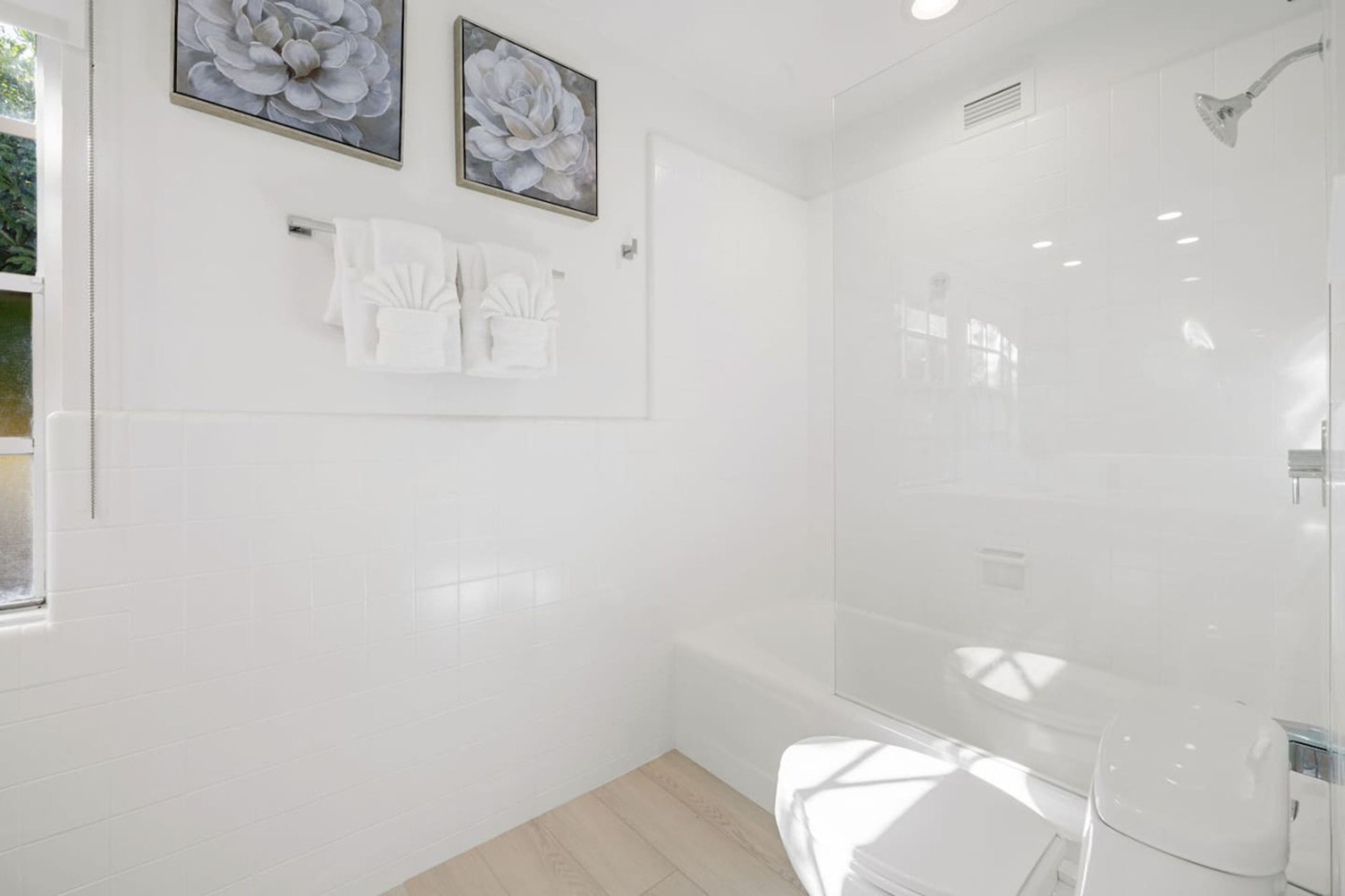 A modern bathroom with white tiles, a glass shower enclosure, and two floral paintings on the wall.