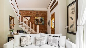 Un canapé blanc orné de coussins à motifs, un escalier avec des rampes en bois, un mur en briques et des œuvres d’art encadrées accrochées aux murs.