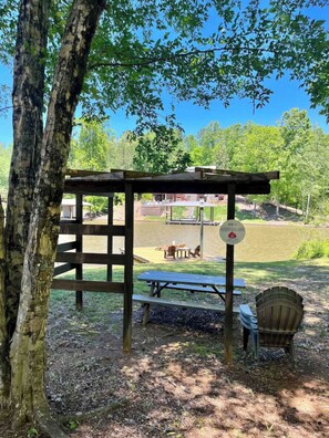 A wooden gazebo with picnic tables and a bench, surrounded by trees and a grassy area.