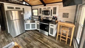 A compact kitchen with a stainless steel refrigerator, a microwave, and a stove. There is a wooden stool and a window providing natural light.