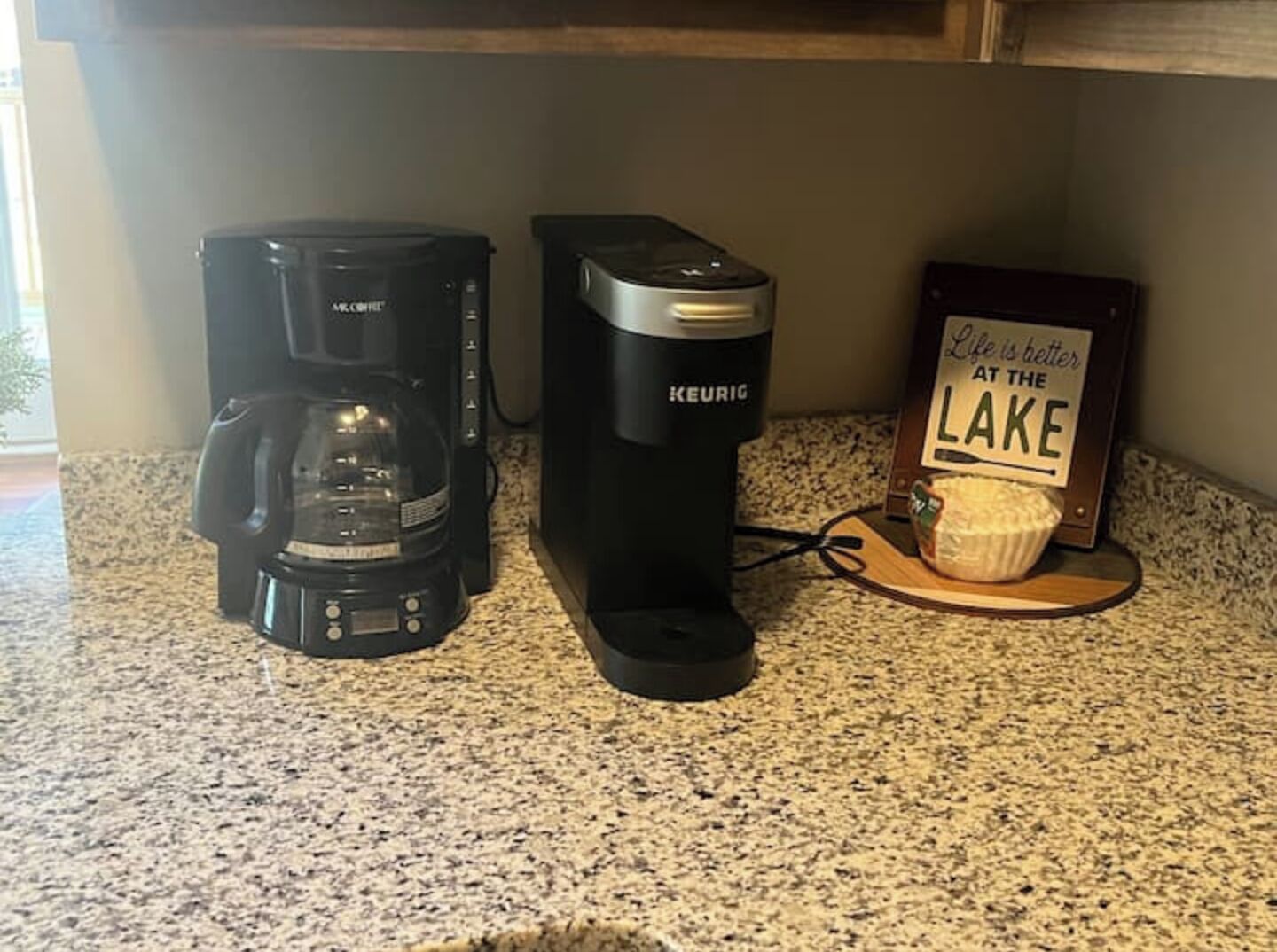 A kitchen counter with two coffee makers, a sign, and a cupcake on a wooden board.