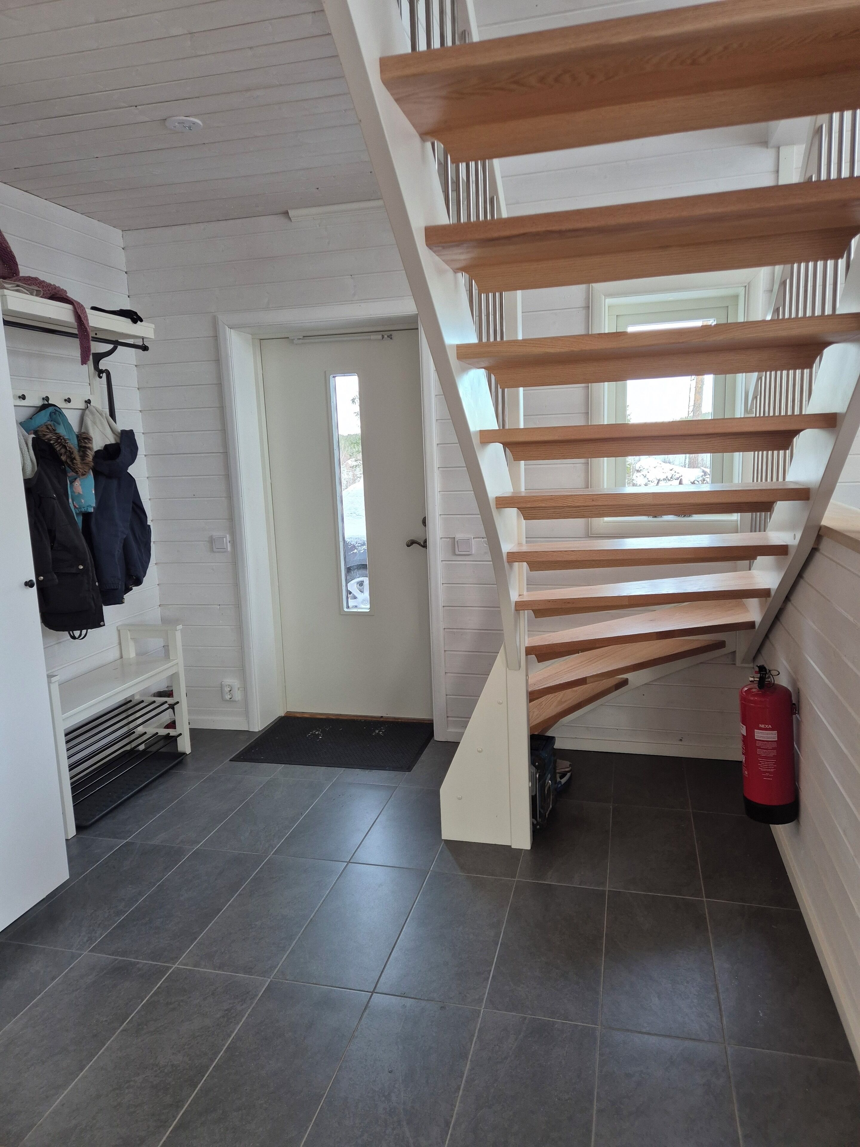 A staircase with wooden steps leading to an upper floor, a coat rack with jackets, a fire extinguisher, and a door leading outside.