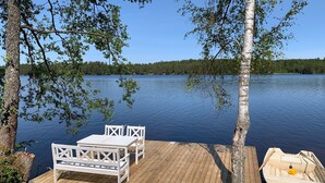A wooden dock with a white table and chairs, a small boat, and a forested shoreline. - Berget Lövanäs 2, Newly built modern house in secluded location with own jetty and boat (Korsberga)