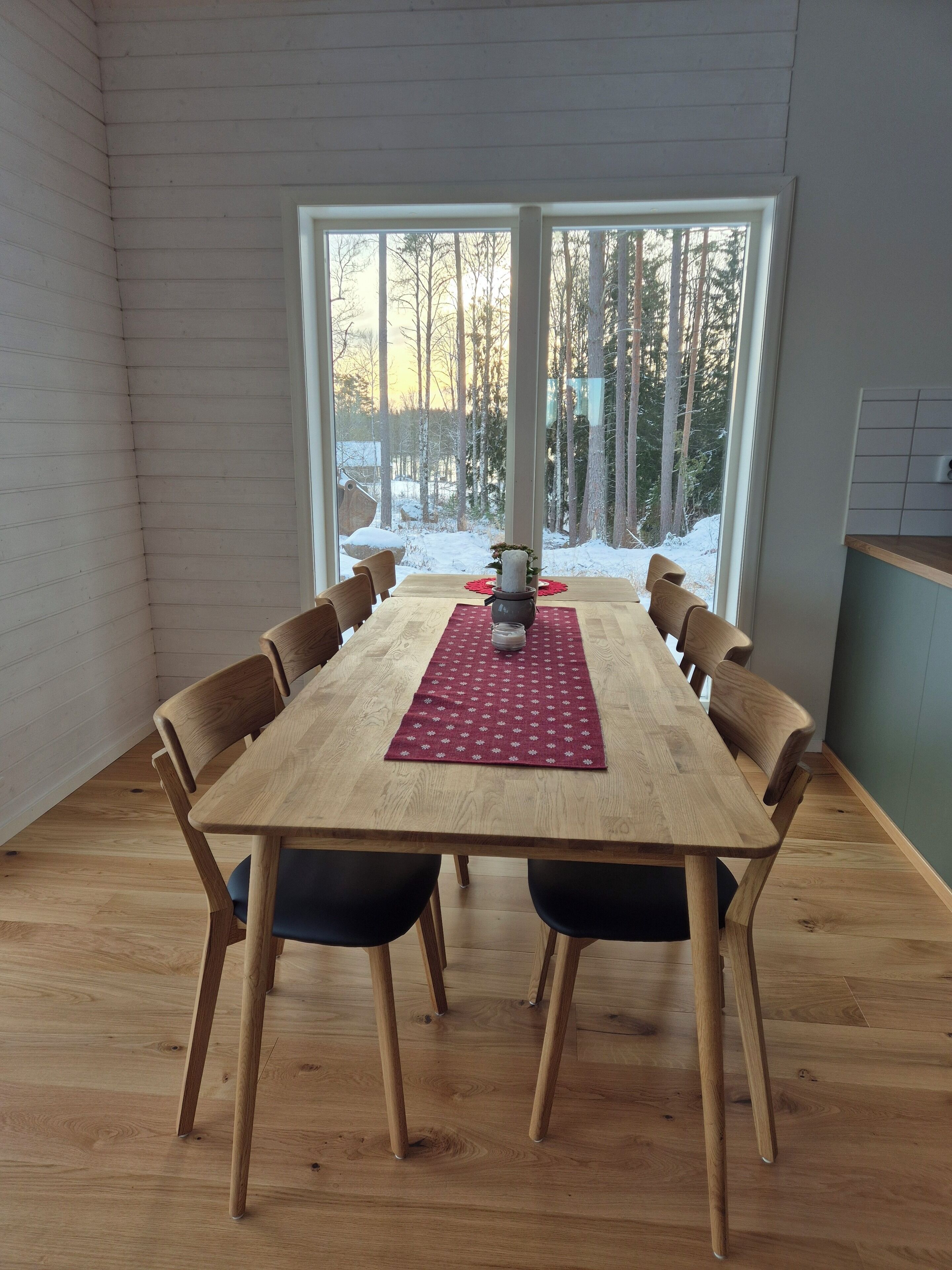 Ein Essbereich mit einem Holztisch, Stühlen, einer roten Tischmatte und Blick auf einen verschneiten Wald durch große Fenster.