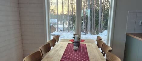 A dining area with a wooden table and chairs, a red placemat, and a view of a snowy forest through large windows.