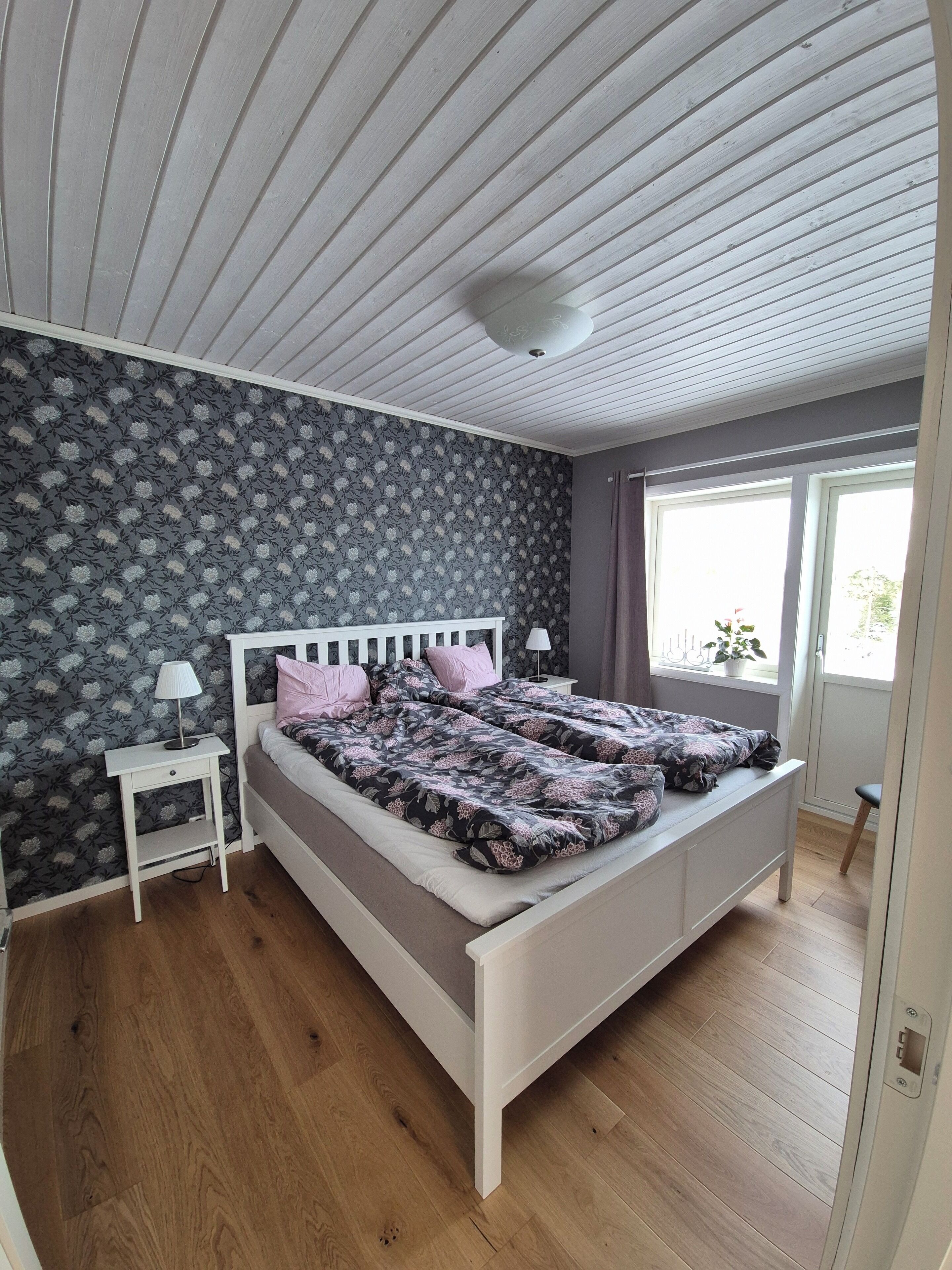 A bedroom with a white bed, floral wallpaper, a nightstand with a lamp, and a window with a view of greenery.
