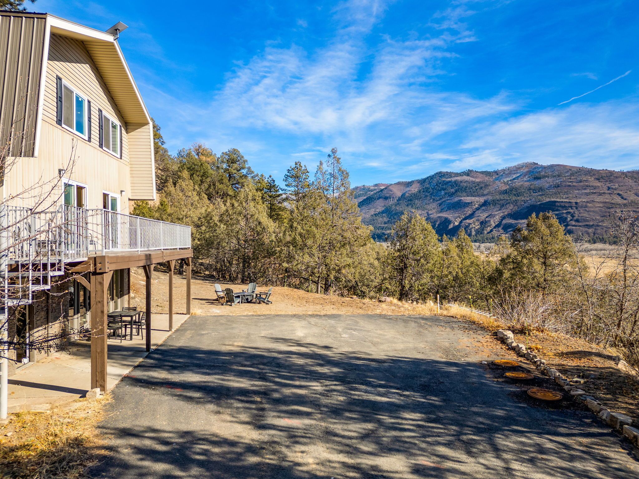 A two-story house with a balcony, a driveway leading to a table and chairs, and a mountainous backdrop.