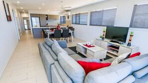 Ein modernes Wohnzimmer mit grauem Sofa, Couchtisch und TV-Möbel. Im Hintergrund ist ein Essbereich mit einer sichtbaren Küche zu erkennen.
