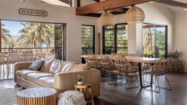 Un grand espace de vie comprenant un canapé, une table à manger et des chaises. Une pancarte indique : « MAISON DE PLAGE ».