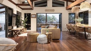 Un vaste espace de vie comprenant un canapé, des poufs ronds, une table à manger avec des chaises en osier et une vue sur l’extérieur.