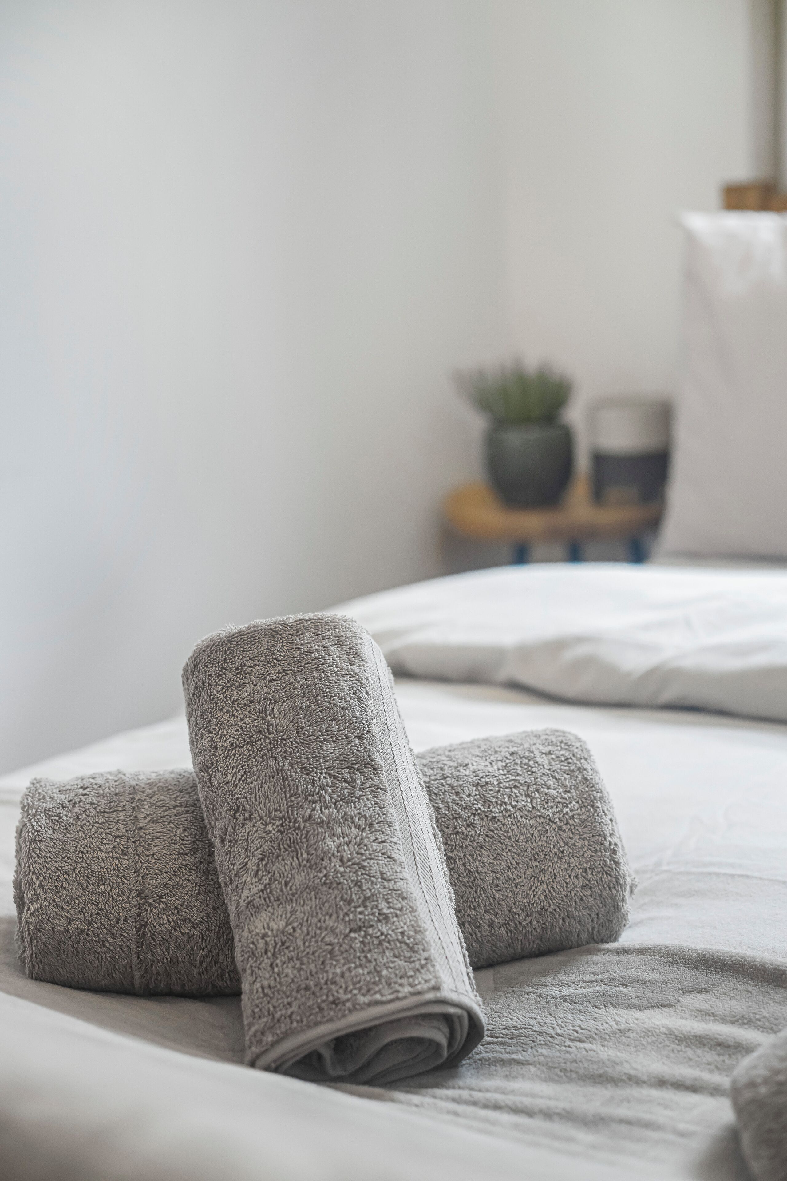 Una cama bien hecha con toallas grises enrolladas y una pequeña planta en maceta sobre una mesita de noche.