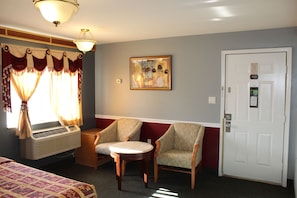 Une chambre d’hôtel comprenant un lit, deux fauteuils, une petite table, une fenêtre avec des rideaux, un climatiseur, une porte avec un numéro et une œuvre d’art accrochée au mur.