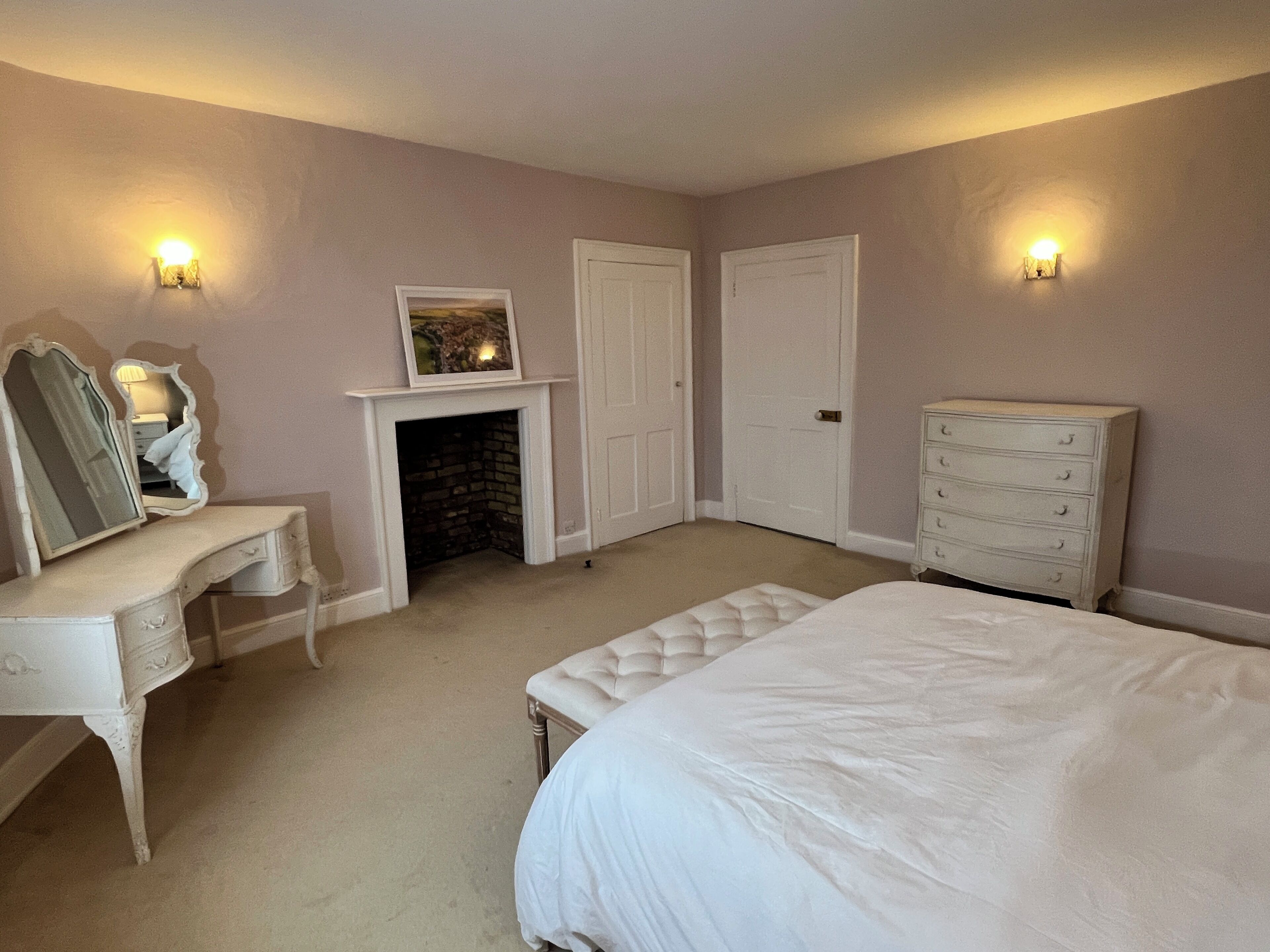 A bedroom with a white bed, dresser, and vanity with a mirror. There is a fireplace, two doors, and wall sconces.
