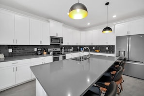 Una cocina moderna con armarios blancos, una isla central, electrodomésticos de acero inoxidable e iluminación colgante.
