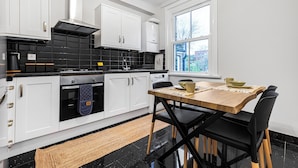 A modern kitchen with white cabinets, a black tiled backsplash, and a wooden dining table with black chairs. - Modern 2 Bedroom Townhouse Flat in King's Cross (London)