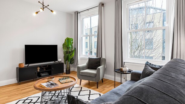 A modern living room with a flat-screen TV, a grey armchair, a black sofa, and a wooden coffee table with chess pieces. - Modern 2 Bedroom Townhouse Flat in King's Cross (London)