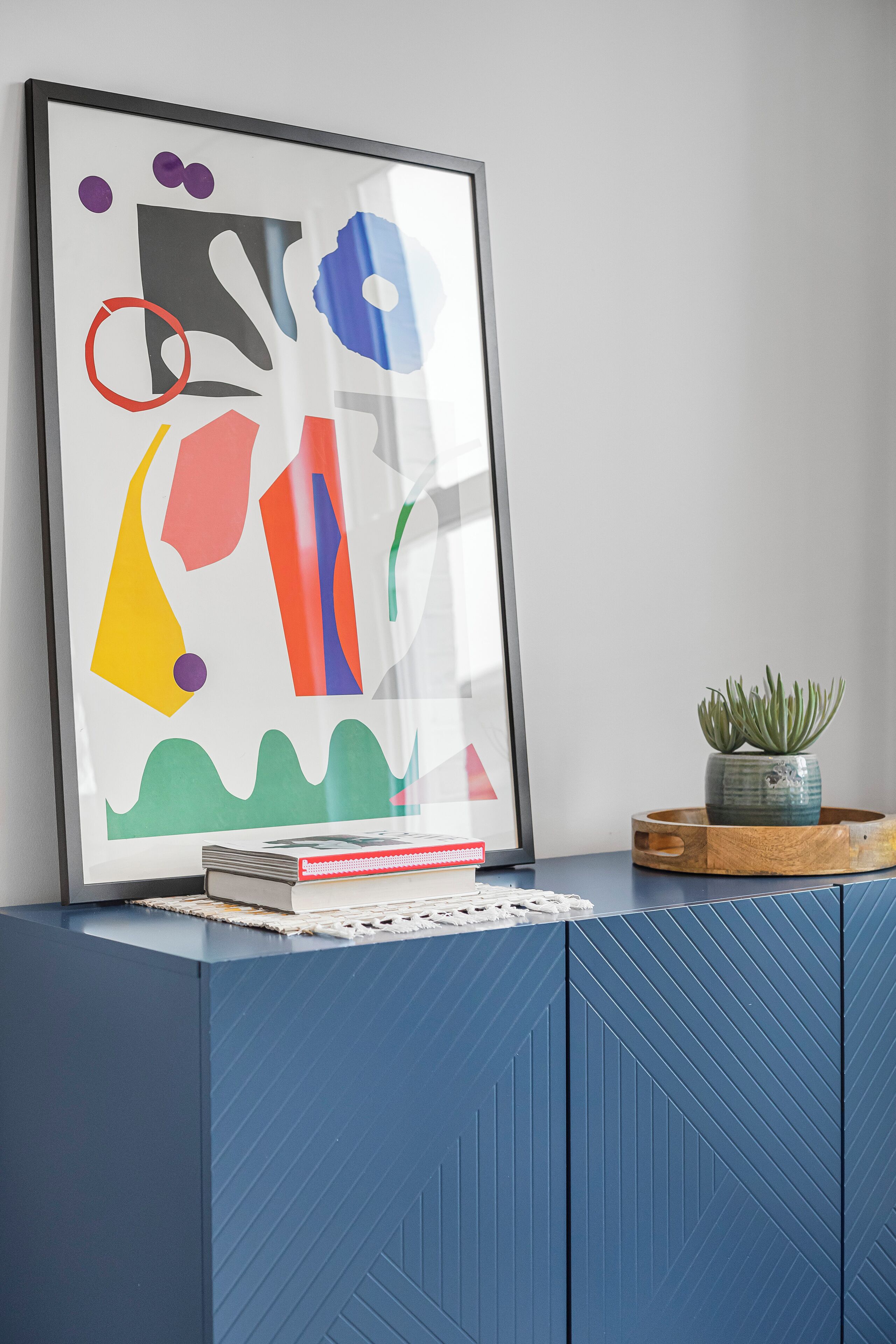 Abstract kunstwerk in een lijst op een blauwe kast, met een kleine plant in een pot ernaast.