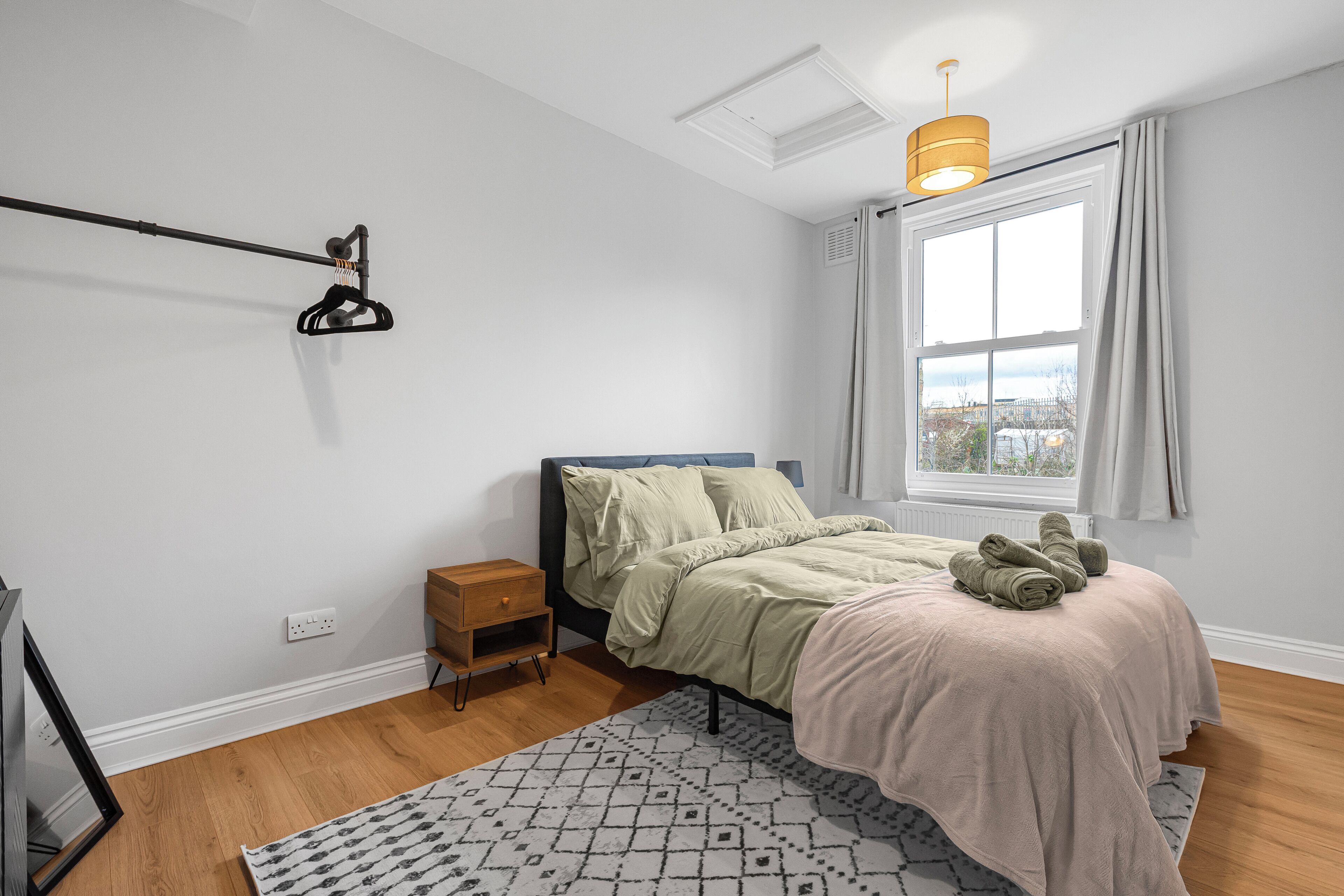 Un dormitorio con cama, una ventana con cortinas, una mesita de noche, una lámpara y vistas a la ciudad.
