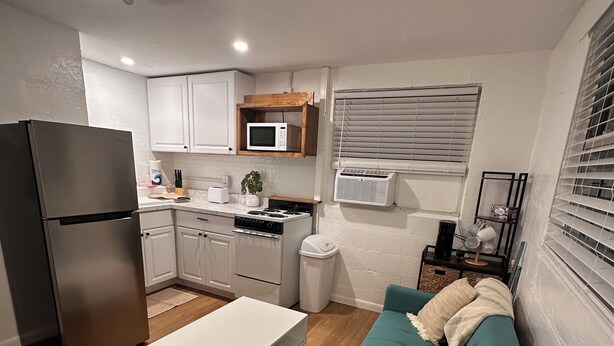 Cocina compacta con refrigerador, microondas, estufa y gabinetes blancos. Un sofá color turquesa con almohadones en la sala, y un ventilador de techo es visible.