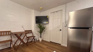 Cocina compacta con refrigerador de acero inoxidable, mesa y sillas de madera, una planta en maceta y una puerta blanca con televisor montado.