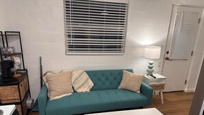 Un sofá azul verdoso con tres almohadones, una mesita de centro blanca, una lámpara y una ventana con persianas.