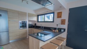 A modern kitchen with wooden cabinets, stainless steel appliances, a black countertop, and wooden stools. - Contemporary family home with outdoor pool and garden (Prinquiau)