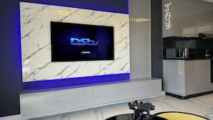 Een moderne woonkamer met een grote flatscreen-televisie bevestigd aan een marmeren muur, een strak keukenhoekje met een koffieapparaat, en een elegante zwarte en gele fauteuil.