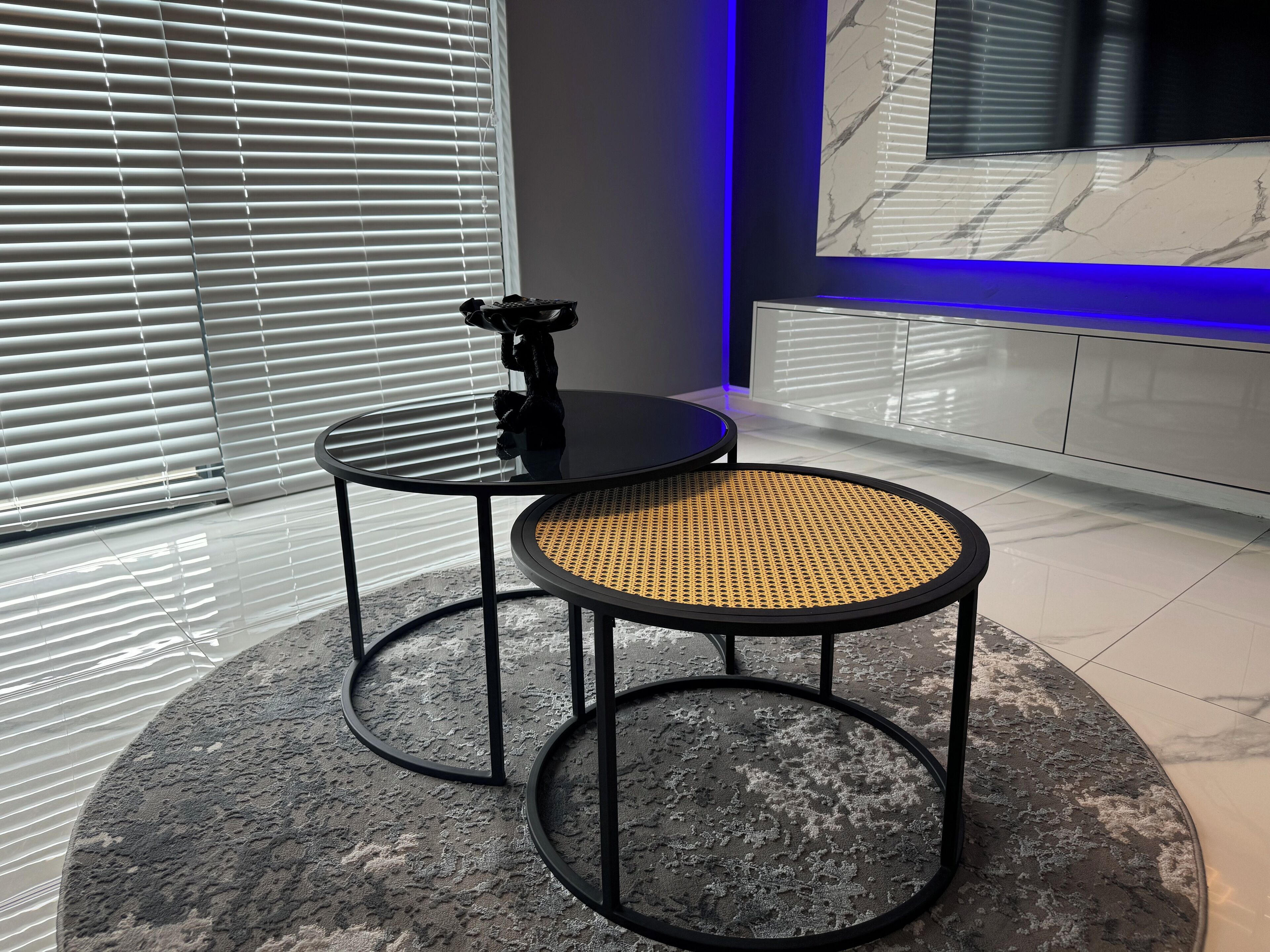 A modern room with two round tables, one with a black surface and the other with a woven seat, and a marble wall with a TV.