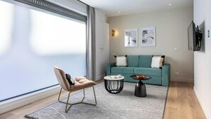 A modern living room with a teal sofa, a wooden floor, a large window, and a minimalist design.