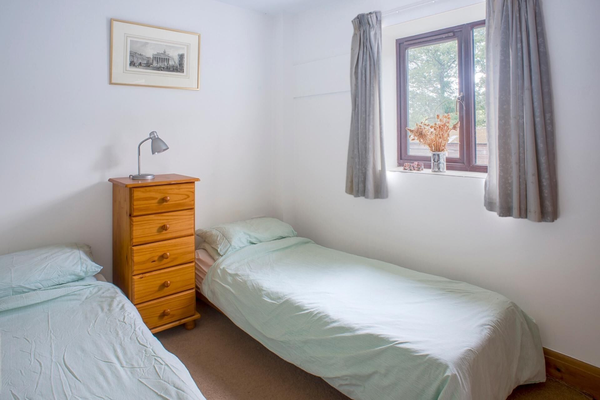 A small, simple room with two beds, a wooden dresser, a desk lamp, a framed picture on the wall, and a window with curtains.