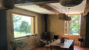Ein rustikaler Essbereich mit einem großen Holztisch und Stühlen, einem fest eingebauten Holzofen und großen Fenstern mit Blick ins Freie.