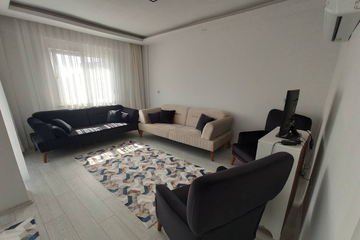 Ein modernes Wohnzimmer mit einer schwarzen Couch, einer beigefarbenen Couch und einem schwarzen Sessel. Auf dem Boden liegt ein Teppich mit Muster, ein Fernseher steht auf einem Schrank und es gibt ein Fenster mit transparenten Vorhängen.