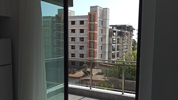 A modern apartment with a large sliding glass door leading to a balcony. The balcony has a metal railing and a view of a construction site with unfinished buildings. - Şahiner Apart Otel (Finike)