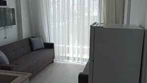 A modern living room with a grey sofa, a flat-screen TV mounted on the wall, a ceiling light fixture, and a large window with sheer curtains. - Şahiner Apart Otel (Finike)