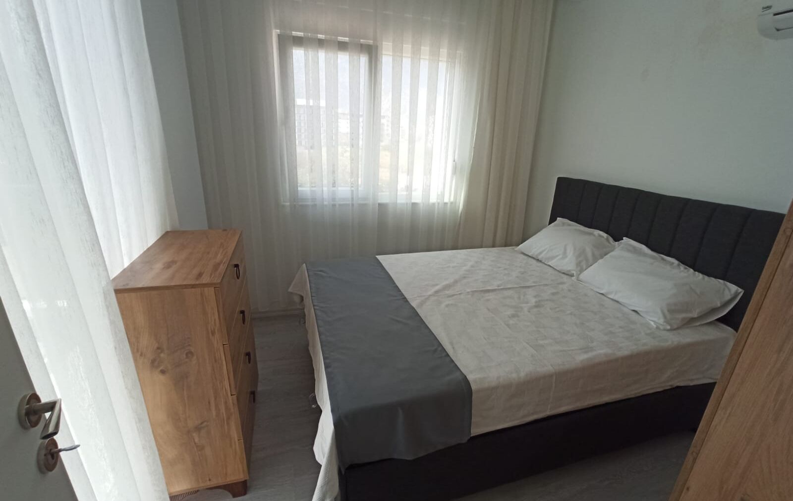 Une chambre à coucher avec un lit, une commode en bois et une fenêtre ornée de rideaux légers.